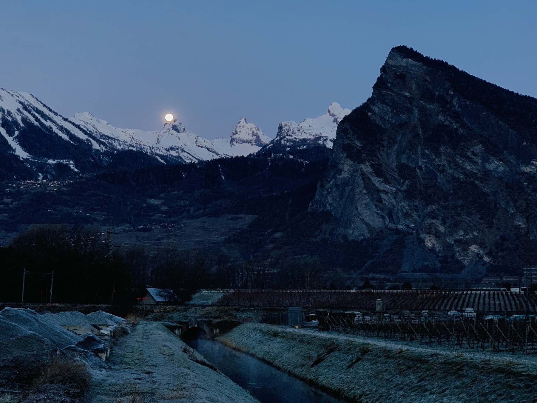 Une chaîne de montagnes éclairée par la lune avec un pic rocheux proéminent sur la droite, qui surplombe un petit canal au premier plan.