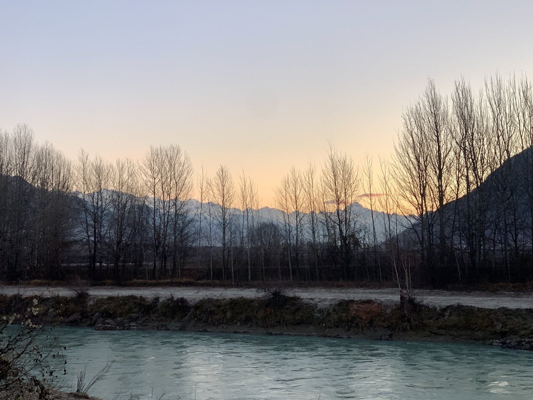 Une rivière au premier plan avec une rangée d'arbres nus et des montagnes lointaines sous un lever de soleil orangé.